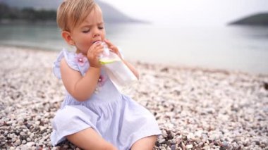 Little girl sits on a pebble beach and drinks water with a lemon from a bottle. High quality 4k footage