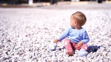 Small child sits on a pebbly beach and plays with a sock taken off his foot. High quality 4k footage