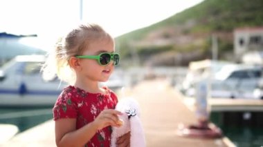 Little girl with a plush hare stands on the pier and eats a bagel. High quality FullHD footage