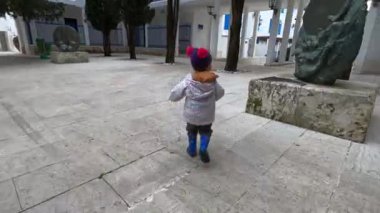 Little girl runs along the tiled yard between buildings, overtaking woman with a stroller. High quality 4k footage