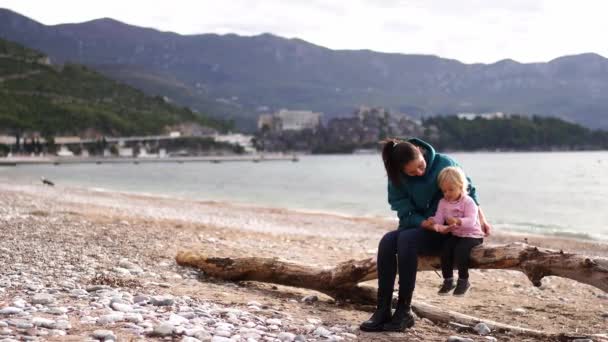 妈妈和一个小女孩坐在海边的卵石沙滩上的一个陷阱上 高质量的4K镜头 — 图库视频影像