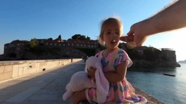 Dad feeds a little girl from a spoon while sitting on the bridge to the island of Sveti Stefan. High quality 4k footage