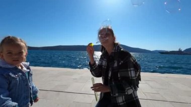 Smiling mother blowing soap bubbles next to a little girl on the seashore. High quality 4k footage