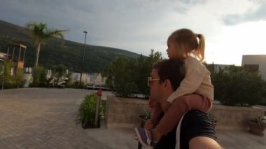 Omuzlarında küçük bir kızla bir tatil köyünün kaldırımlı caddelerinde yürüyen bir baba. Yüksek kalite 4k görüntü