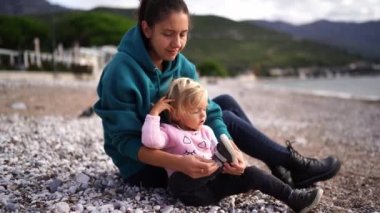 Annem sahilde oturan küçük bir kızın ayakkabılarından çakıl taşlarını sallıyor. Yüksek kalite 4k görüntü