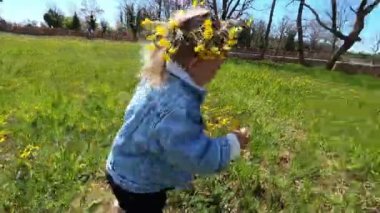 Little girl in a wreath of yellow dandelions runs along a green lawn. High quality 4k footage