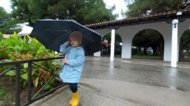 Little girl in a raincoat and rubber boots walks under an umbrella along a path in the park. High quality 4k footage