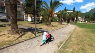 Panama giymiş küçük bir kız, parktaki yolda elinde oyuncakla bir bebek arabasını yuvarlıyor. Yüksek kalite 4k görüntü