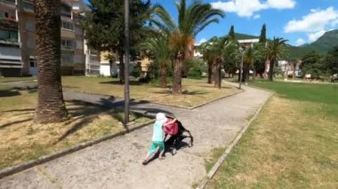 Little girl in a panama rolls a stroller with a toy along the path in the park. High quality 4k footage