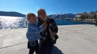 Mother blowing soap bubbles next to a smiling little girl on the seashore. High quality 4k footage