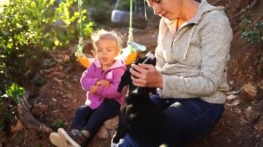 Mom and little girl are sitting on the ground in the garden and stroking a black puppy. High quality 4k footage