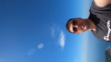 Young man takes off his sunglasses while standing on top of a mountain above the bay. High quality 4k footage