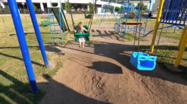 Little girl swings on a chain swing in a playground near a road with moving cars. High quality 4k footage
