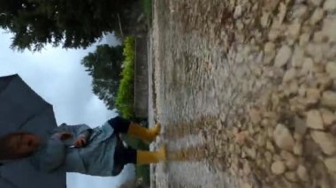 Little girl with an umbrella jumps through the puddles on the path in the park in the rain. High quality 4k footage