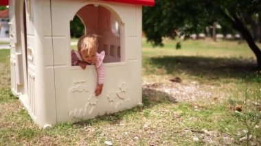 Little girl looks out of the window of a toy house on the playground, and then leaves it. High quality 4k footage