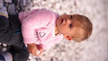 Little girl sits on a pebbly beach with a pebble in her hands and looks back. High quality 4k footage