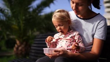 Little girl eats porridge with a spoon, sitting in her mom arms in the park. High quality 4k footage