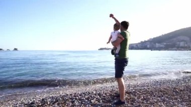 Dad with a small child in his arms throws pebbles into the sea while standing on a pebble beach. High quality 4k footage