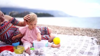 Mom strokes the head of a little girl sitting on the beach and playing with pebbles and toys. High quality 4k footage