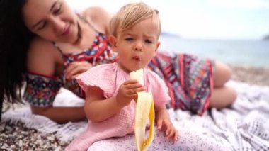 Mom lies on a blanket on the beach next to a little girl eating a banana. High quality 4k footage
