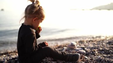 Little girl nibbles an apple sitting on a pebble beach and looks at the sea. High quality 4k footage