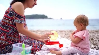 Mom and little girl play with toy dishes while sitting on the beach. High quality 4k footage