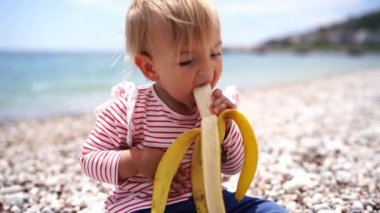 Little girl peels a banana and eats it while sitting on the beach. High quality 4k footage