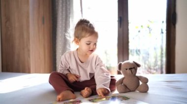 Little girl showing picture book to teddy bear while sitting on bed. High quality 4k footage