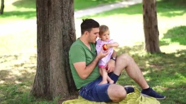 Dad gives a big apple to a little girl sitting in his arms under a tree on a bedspread. High quality 4k footage