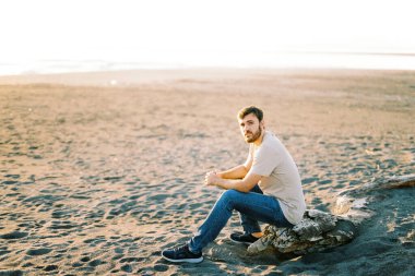 Young man in jeans sitting on a snag on a sandy beach. High quality photo