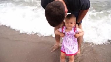 Dad holds a little girl by the shoulders while standing on the sand in the surf. High quality 4k footage