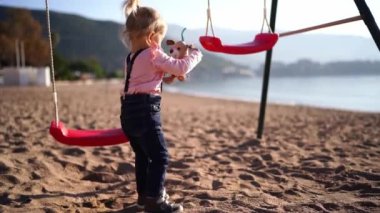 Little girl shakes a plush dog while standing on the beach near the swing. High quality 4k footage