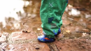 Small child in overalls and rubber boots steps on a puddle. High quality 4k footage