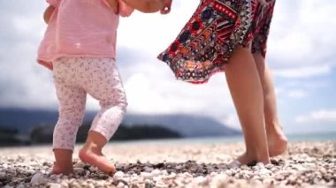 Little girl learning to walk on a pebbly beach holding her mother hand. High quality FullHD footage