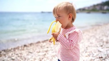 Little girl eating a banana from two hands while standing on the beach. High quality 4k footage