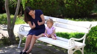 Mom claps her hands for a little girl sitting with her on a park bench. High quality 4k footage