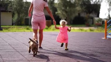 Dog runs after mom and little girl walking on the playground. High quality 4k footage