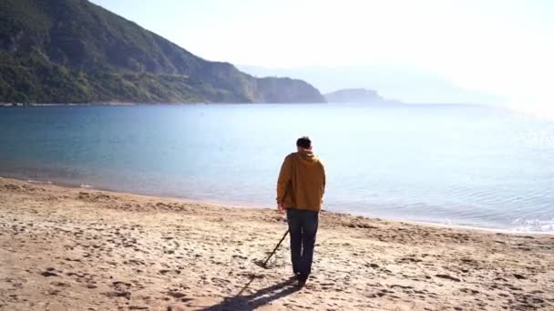 一个带着金属探测器的人走在海边靠近大海的地方 后视镜高质量的4K镜头 — 图库视频影像