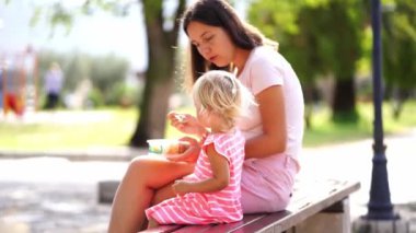Mom feeds a little girl lunch from a spoon, sitting on a bench. High quality 4k footage