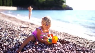 Little girl puts pebbles into toy buckets while sitting on the beach. High quality 4k footage