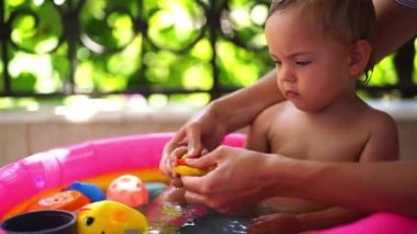 Mom teaches a little girl to splash water from a rubber duck while sitting in an inflatable pool. High quality 4k footage