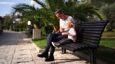 Mom feeds a little girl from a spoon sitting on a bench in the park. High quality 4k footage