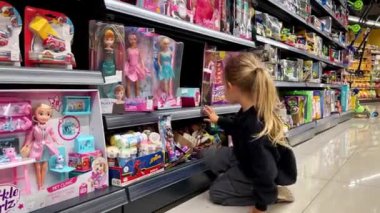Küçük kız bir süpermarketin zemininde oturuyor ve raflardaki bebekleri seçiyor. Yüksek kalite 4k görüntü