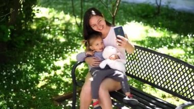 Anne ve küçük bir kız bahçede bir bankta otururken akıllı telefondan selfie çekiyorlar. Yüksek kalite 4k görüntü
