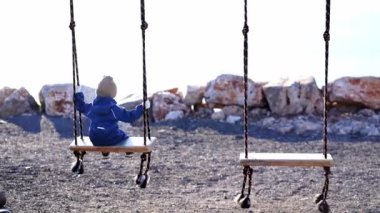Tulumlu ve eldivenli küçük bir çocuk çakıl taşlı plajda sallanıyor. Arkadan bak. Yüksek kalite 4k görüntü