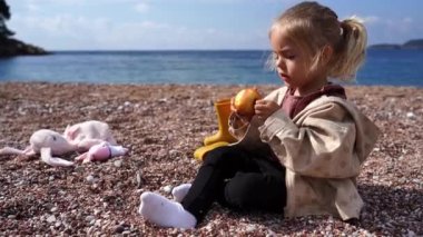 Küçük kız bir çakıl taşı kumsalında ceketiyle oturuyor ve bir elma yiyor. Yüksek kalite 4k görüntü