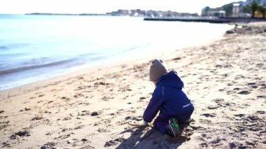 Tulumlu ve eldivenli küçük kız deniz kenarındaki kumlu sahilde oynuyor. Yüksek kalite 4k görüntü