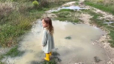 Lastik çizmeli küçük bir kız derin bir su birikintisinde yürüyor, elbisesinin kenarını kaldırıyor. Yüksek kalite 4k görüntü