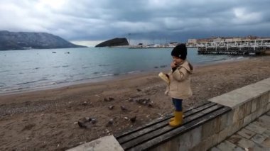 Lastik çizmeli küçük bir kız denizin yanındaki taş bir çitin üzerinde duruyor ve bir somun ekmek yiyor. Yüksek kalite 4k görüntü