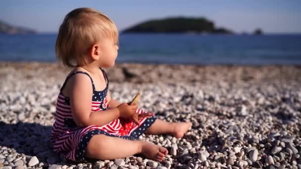 小さな女の子は小石のビーチに座ってパンケーキを食べ 海を見ています 高品質4K映像 — ストック動画